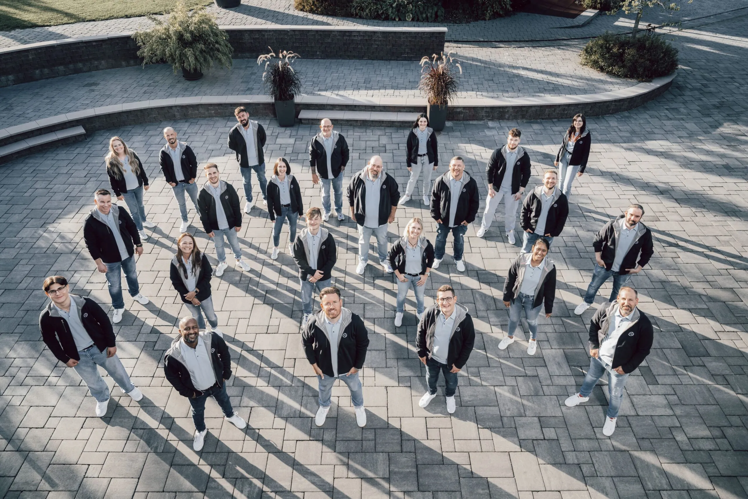 Foto des Teams der Neo One AG (Drohnenaufnahme). Alle Teammitglieder schauen in die Kamera, tragen Jeans, dunkle Jacke, ein hellgraues Hemd und weiße Turnschuhe.